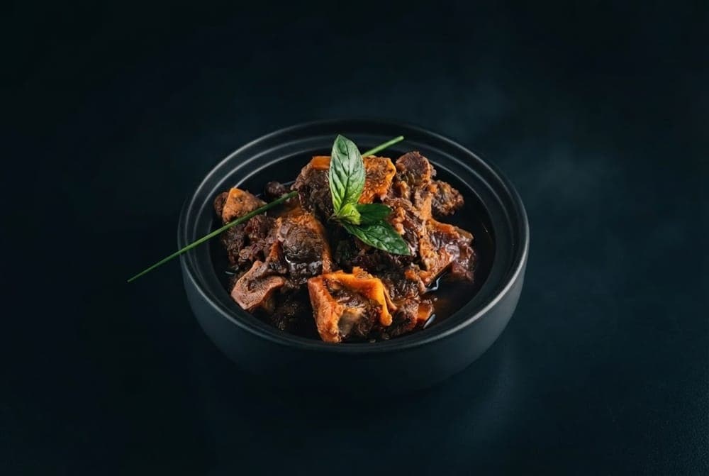 Braised oxtail in a dark, glossy gravy, garnished with fresh herbs in a black bowl