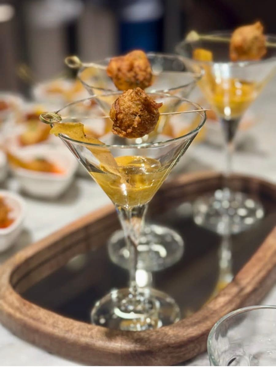 Martini glasses of a golden drink, each topped with a fritter on a skewer