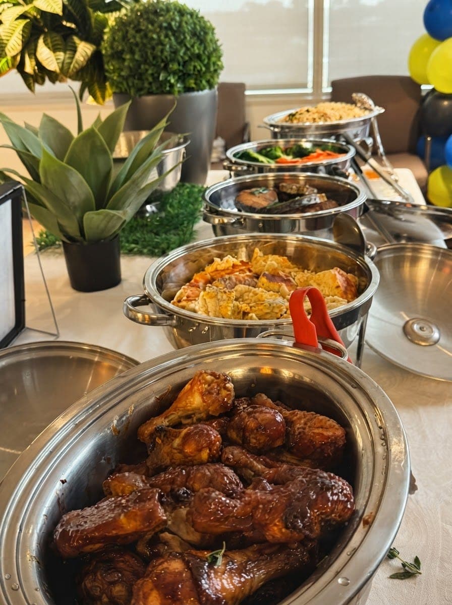 A chafing dish of glazed chicken beside trays of rice and stews on a buffet